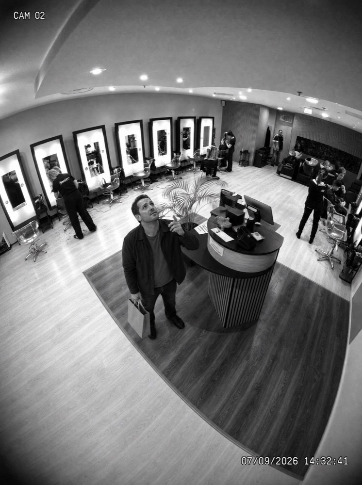 Image de caméra de surveillance en noir et blanc : dans un salon de coiffure, un client lève les yeux vers l’objectif, image générée par IA