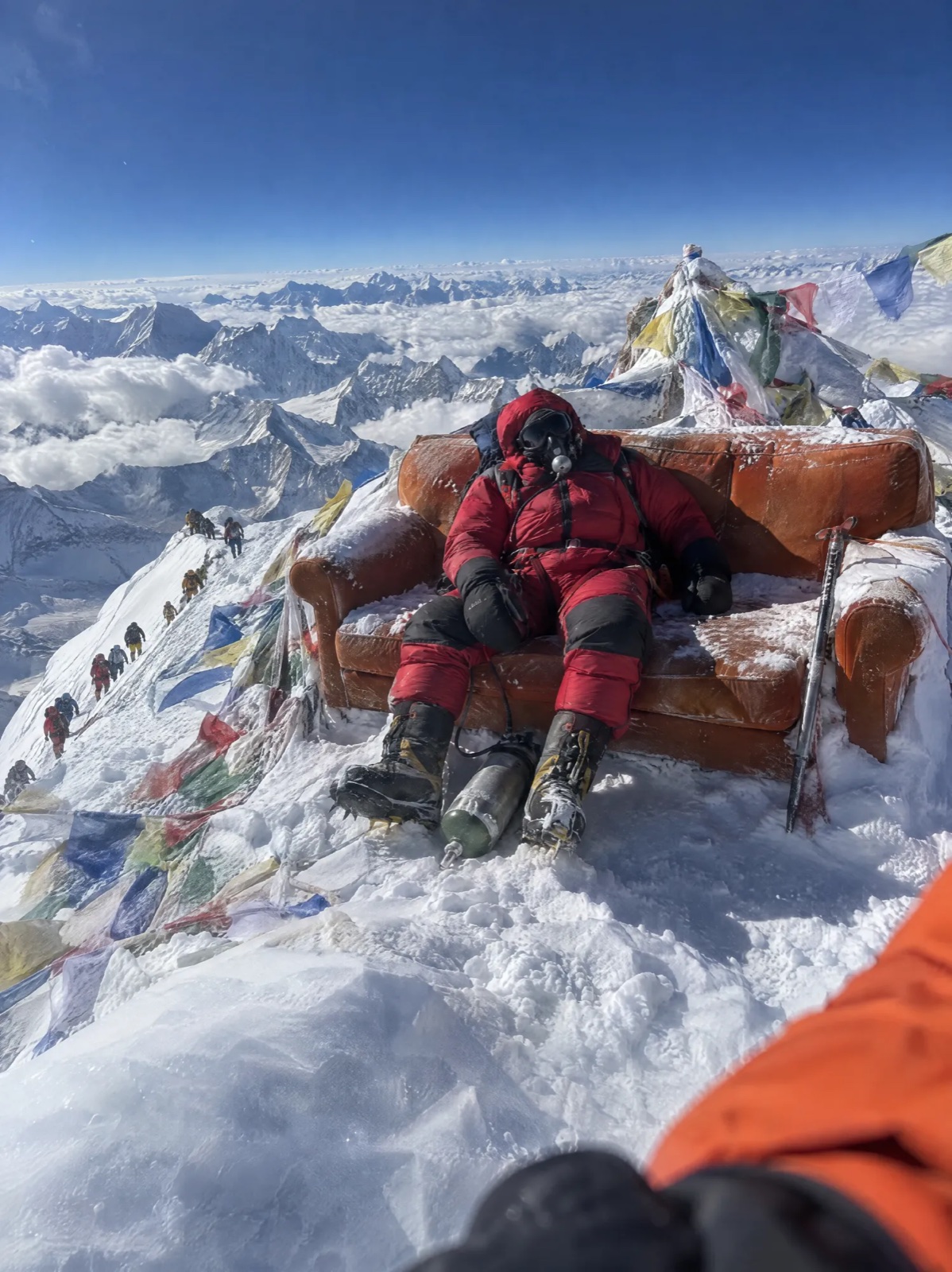 Un alpiniste se repose sur un canapé orange posé au sommet de l’Everest, au milieu des drapeaux de prière, image générée par IA